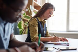 Female Teacher Writing in Notebook Sitting at Desk during English Lesson