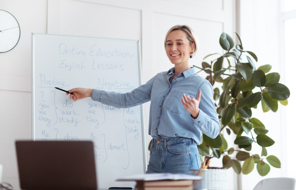Joyful young teacher standing near blackboard, explaining grammar, giving online lesson to students