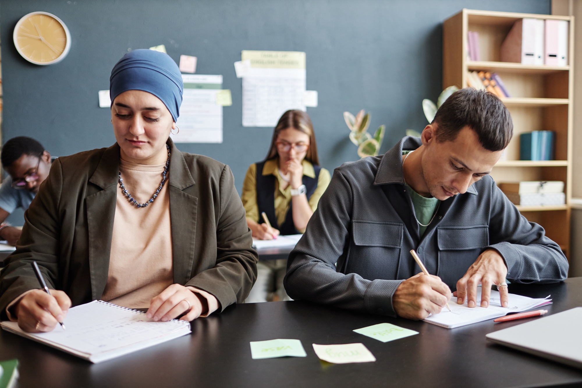 Muslim Woman Writing in Notebook Sitting at desk next to Man during Lesson in Classroom