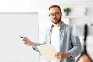 Young male teacher pointing at blackboard with English grammar rules, conducting online lesson from